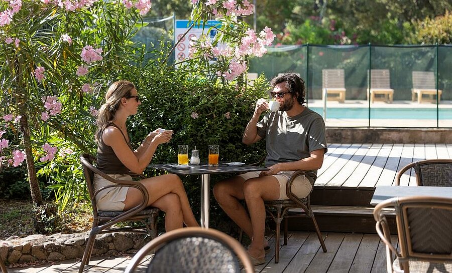 Couple en terrasse ombragée avec boissons, près de la piscine d’un camping en Corse